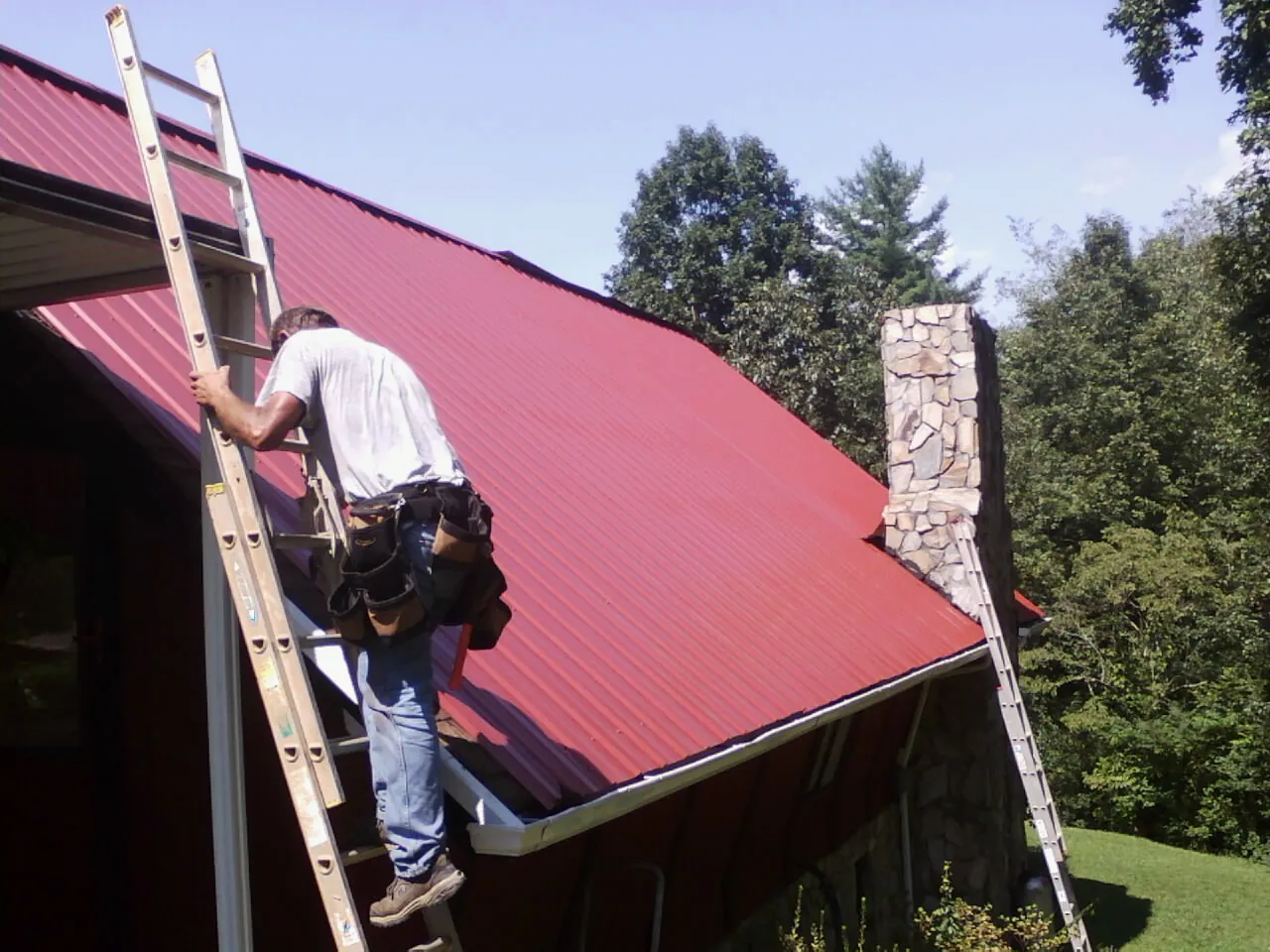 Roofer on a ladder beside a red metal roof and stone chimney — Soffit & Fascia Repair by Roofing Brothers in Grand River